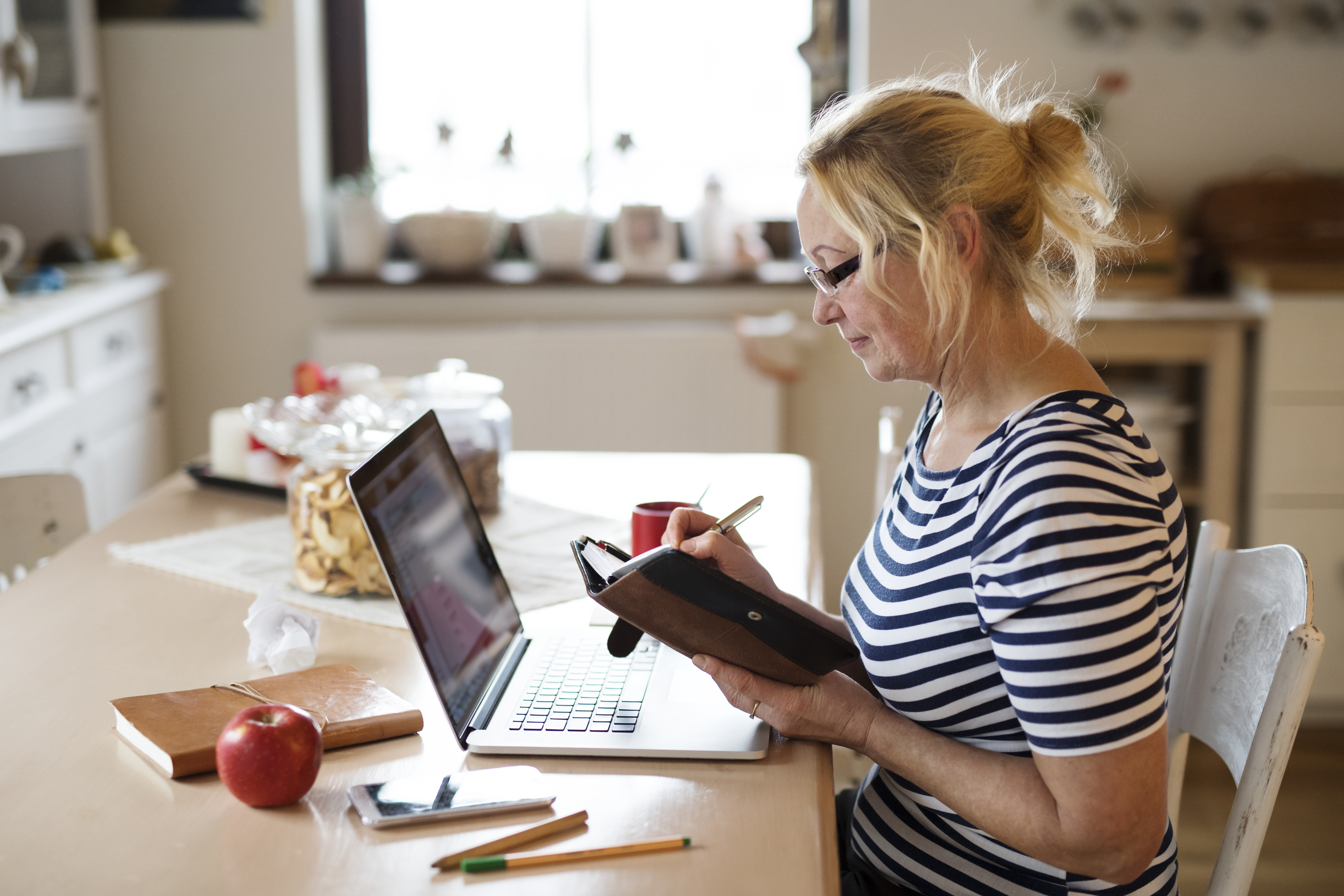 Blonde Mature Woman Looking At Paper Diary With Laptop